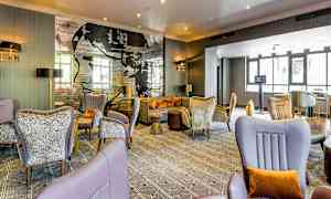 Ett modernt hotellrum med bekväma sittmöbler, eleganta dekorationer och en väggklistertapet med text och konst. Stora fönster släpper in naturligt ljus. (AI-genererad text)