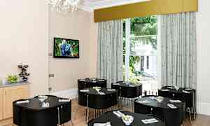 Ett ljust rum med flera svarta mörka bord och stolar samt en stor fönster mot grönska. (AI-genererad text)