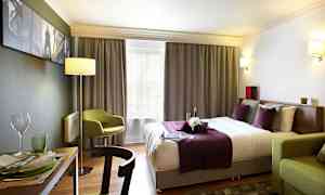 Ett hotellrum med en dubbelsäng klädd i lila och vita sängkläder, en grön stol och ett matbord med porslin. (AI-genererad text)