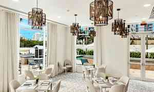 Ett ljust och stilrent inrett rum med vita möbler, stora fönster och hängande lampor. Utsikten visar en pool och omgivande byggnader. (AI-genererad text)