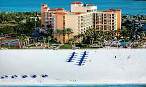 Ett hotellkomplex vid stranden, med blå parasoller och solstolar på den vita sanden, och en bakgrund av havet. (AI-genererad text)