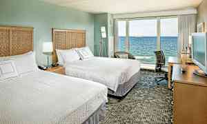 Hotellrum med två dubbelsängar, en TV på ett skrivbord och en stor glasvägg med utsikt över havet. (AI-genererad text)