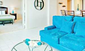 En ljus och rymlig interiör med ett vardagsrum och ett sovrum. I förgrunden syns en blå soffa samt ett glasbord med dekorationer. Sovrummet har en säng och en nattlampa. (AI-genererad text)