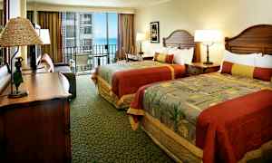 Hotellrum med två bäddar som har färgglada sängöverkast, ett skrivbord, och en öppen balkong med utsikt över havet. (AI-genererad text)