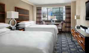 Ett hotellrum med två dubbelsängar klädda i vita sängkläder, ett bord med stolar nära ett fönster samt en byrå med en TV ovanpå. (AI-genererad text)