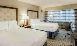 Ett hotellrum med två dubbelsängar, en lampa och ett skrivbord bredvid en stol. (AI-genererad text)