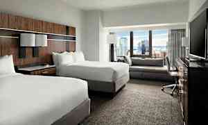 Ett hotellrum med två dubbelsängar, en sittgrupp, ett skrivbord och en TV, med utsikt över en stad genom fönstret. (AI-genererad text)