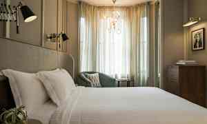 Ett elegant sovrum med en stor säng täckt av vita lakan, en grön fåtölj, och mjuka gardiner som släpper in ljus. (AI-genererad text)