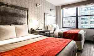 Ett hotellrum med två sängar med vita sängkläder och orangea överkast. Det finns ett fönster med utsikt över en stad. (AI-genererad text)