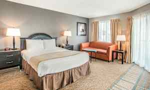 Ett hotellrum med en dubbelsäng, en nattduksbord på vardera sidan, en liten soffa, soffbord och tre lampor. Fönster med gardiner syns i bakgrunden. (AI-genererad text)