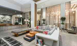 Ett modernt vardagsrum med stora fönster, grå soffa med färgglada kuddar och interiördetaljer som planterade krukväxter och faktaböcker. (AI-genererad text)