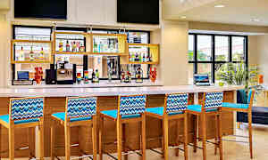 En bar med höga stolar i trä och blåa ryggstöd, en bänkskiva med diverse drycker och en kaffemaskin. (AI-genererad text)