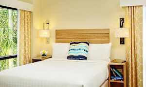 Ett sovrum med en säng bäddad med vitt sänglinne, en dekorativ kudde och nattygsbord. Gardiner med gult mönster inramar fönstren. (AI-genererad text)