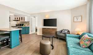 Ett vardagsrum med en soffa, fåtölj, och en TV på väggen. En köksdel med blå skåp syns till vänster. (AI-genererad text)