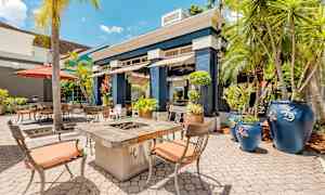 En solig uteplats med bord och stolar, en eldstad och dekorativa blå krukor omgivna av palmträd. (AI-genererad text)