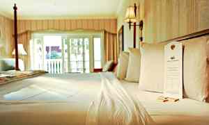 Säng i ett hotellrum med vita lakan och kuddar, samt tillhörande inredning och ljusinsläpp från en balkongdörr. (AI-genererad text)