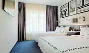 Ett hotellrum med två enkelsängar med vita lakan och dekorerade överkast. Rummet har mörklila gardiner, en väggmonterad TV och inramade bilder på väggen över sänggaveln. (AI-genererad text)