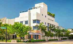 En vit byggnad i modern stil med blå fönsteromgivningar, omgiven av träd och palmer. (AI-genererad text)