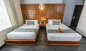 Bild av ett hotellrum med två enkelsängar med vita sängkläder och mönstrade överkast. Rummet har en träpanel på väggen och ett nattduksbord med en blomma mellan sängarna. (AI-genererad text)