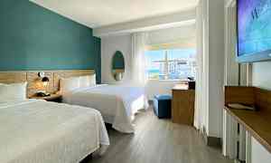 Ett modernt hotellrum med två dubbelsängar, ett skrivbord och ett stort fönster som släpper in naturligt ljus. (AI-genererad text)