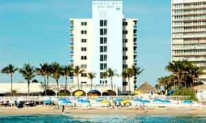 Ett hotell med palmträd framför, bredvid stranden med parasoller och människor på sanden. (AI-genererad text)