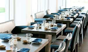 Ett elegant restaurangområde med dukade bord, blå servetter och glas. Ljusa fönster i bakgrunden. (AI-genererad text)