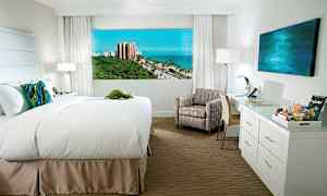 Hotellrum med en bred säng, stol, skrivbord och utsikt över havet och grönska utanför. (AI-genererad text)