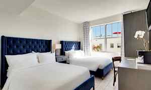 Ett hotellrum med två sängar med blåa sänggavlar, ett nattduksbord med lampa mellan sängarna och ett skrivbord med en blomma och kaffemaskin. (AI-genererad text)