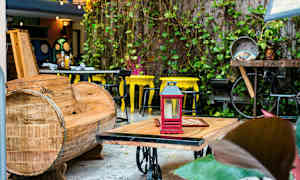 En utomhus plats med en träsoffa, ett bord med en röd lykta ovanpå och gula stolar i bakgrunden. Växter pryder omgivningen. (AI-genererad text)