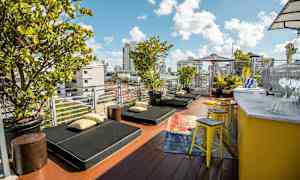 En terrass med solstolar, kuddar och gröna växter. Det finns en gul bardisk med stolar vid sidan. (AI-genererad text)