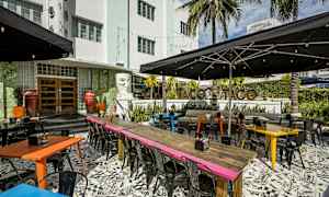En restaurangutomhus med färgglada bord och stolar i en livfull miljö. Bakgrunden visar byggnaden och tropiska palmer. (AI-genererad text)