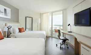 Ett modernt hotellrum med två stora sängar, ett skrivbord med en stol, och en balkong med utsikt över havet. (AI-genererad text)