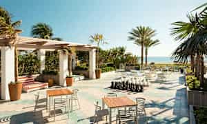 En utomhusterrass med bord och stolar, en schackbräda och utsikt över stranden och havet, omgiven av palmer. (AI-genererad text)