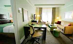 Ett hotellrum med ljus inredning, inklusive en säng, skrivbord, soffa och TV. (AI-genererad text)