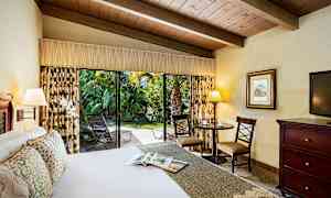 Ett hotellrum med en stor säng, ett bord med stolar och en öppen skjutdörr som leder till en grönskande utomhusmiljö. (AI-genererad text)