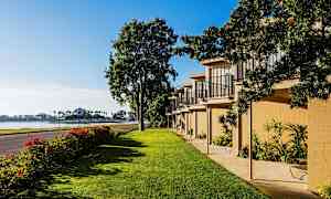 Bilden visar ett grönskande område med gräsmatta, en rad byggnader med balkonger och en väg som löper bredvid. I bakgrunden syns vattnet och palmer. (AI-genererad text)