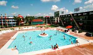 Utomhuspool med solstolar runt om och människor som badar. I bakgrunden syns byggnader med sporttema och en stor dekorativ artefakt som liknar en amerikansk fotboll samt palmer. (AI-genererad text)