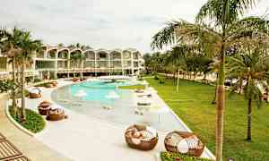 En utomhuspool vid ett modernt hotell omgivet av palmer och grönska. Solstolar och skuggande parasoller finns vid poolen. (AI-genererad text)