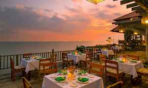 En utomhusrestaurang med dukade bord på en träterrass vid havet under en färgglad solnedgång. (AI-genererad text)