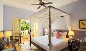 Ett sovrum med en himmelssäng, ljust inrett med vita och rosa kuddar. Ett bord med en lampa står bredvid sängen, och en dörr leder ut till en terrass med grönska. (AI-genererad text)