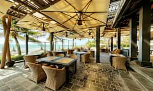 En utomhusrestaurang med korgmöbler och parasoller, belägen vid stranden med utsikt över havet. (AI-genererad text)