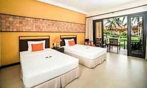 Två enkelsängar med vita sängkläder och dekorativa orange kuddar, ett nattduksbord mellan dem med lampa, telefon och dekorationer. Det finns stora fönster med utsikt mot trädgården. (AI-genererad text)