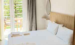 Ett ljust och rymligt sovrum med en dubbelsäng, vita sängkläder och handdukar. Ett stort glasfönster leder ut till en balkong med trämöbler och utsikt över grönska. (AI-genererad text)