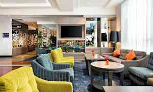 En hotellobby med bekväma sittplatser, flera soffor och stolar i olika färger, samt bord med blommor. En TV är monterad på väggen och det finns dekorativa föremål på hyllorna. (AI-genererad text)