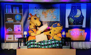 Två tecknade figurerdjur, en giraff och en björn, sitter i en säng med täcke och scenrekvisita synliga i bakgrunden. (AI-genererad text)