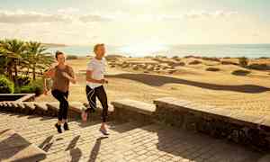 Två personer joggar på en stenlagd gångväg nära sanddynor och palmer vid kusten. (AI-genererad text)