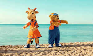 Två maskotar, en giraff och en björn, står på en strand med havet i bakgrunden. (AI-genererad text)