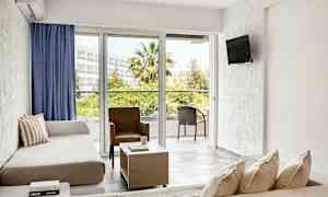 Ett hotellrum med soffa, fåtölj och bord, med utsikt mot en balkong och grönska. (AI-genererad text)