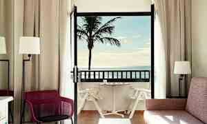Ett hotellrum med balkong där man ser en palm och havsutsikt. (AI-genererad text)