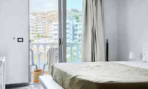 Utsikt från ett hotellrum, med säng och en öppen balkongdörr. Balkong med stolar och utsikt över byggnader. (AI-genererad text)